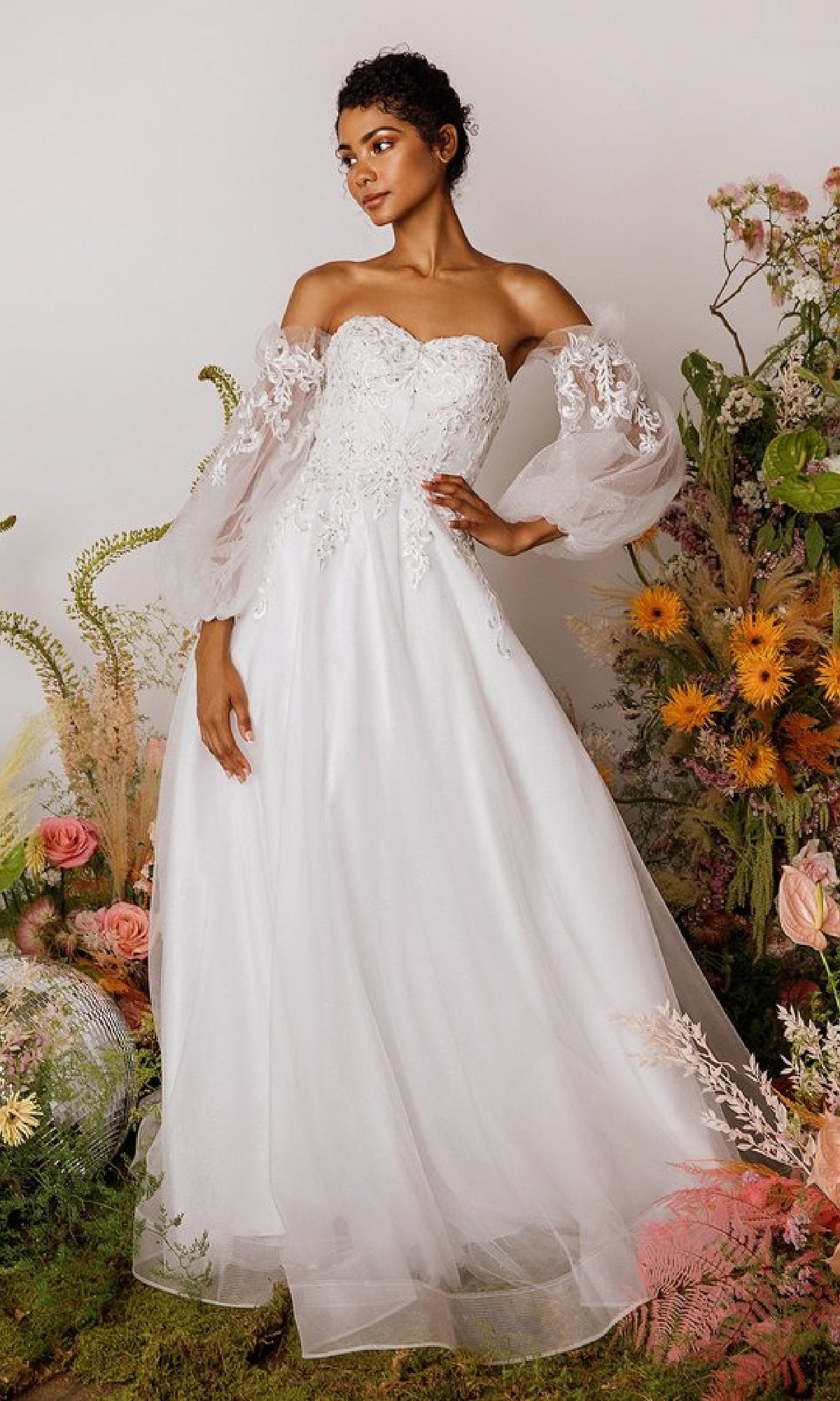 A woman stands among assorted flowers and greenery wearing the Eureka 9505 Long Sleeve Strapless Long Prom Gown, featuring an embroidered sweetheart bodice and sheer lace sleeves.